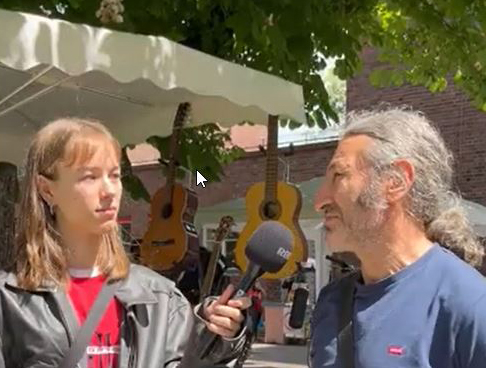 Eine Frau und ein Mann. Die Frau hält ein Mikrofon in der Hand und interviewt den Mann. Im Hintergrund ist ein Flohmarktstand zu sehen, der Instrumente, unterandrem Gitarren, verkauft.